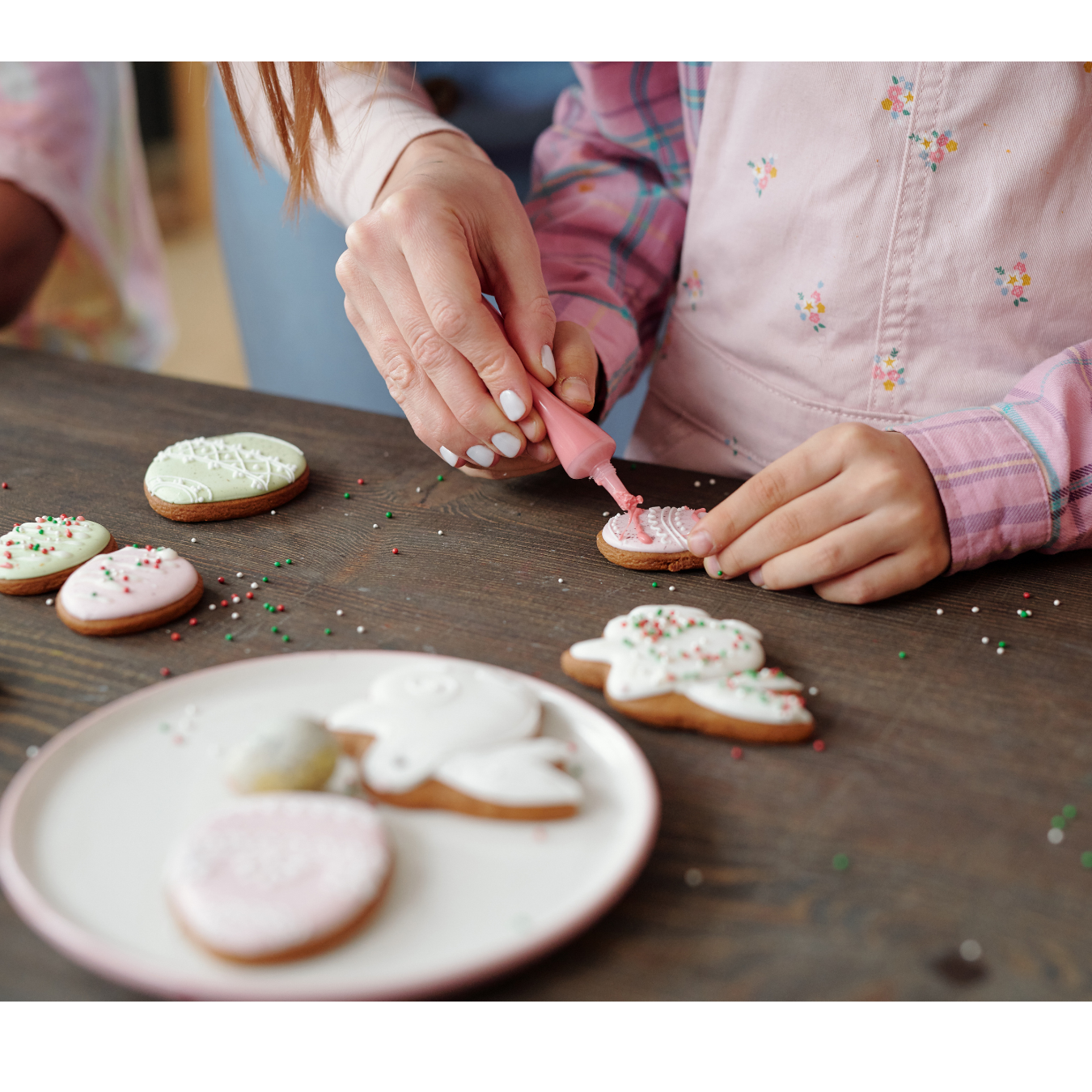 A person using a pink icing bag to decorate a heart-shaped cookie on a wooden table with other decorated treats.