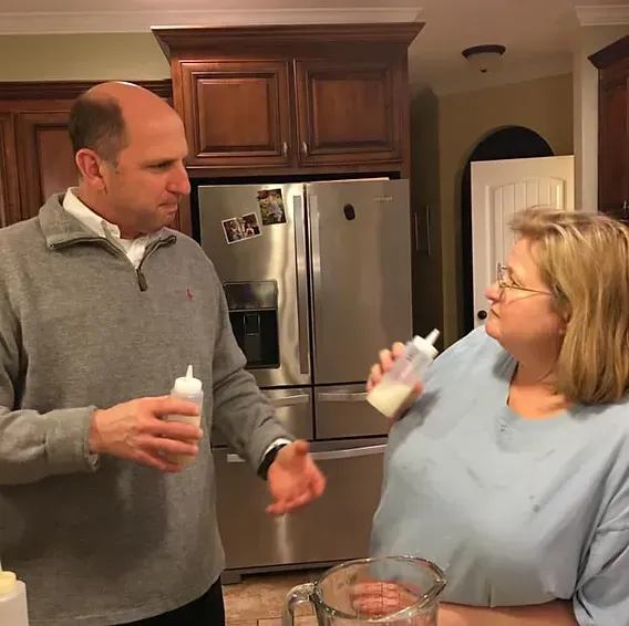 A person in a gray sweater and another in a blue shirt stand in a kitchen, both holding small squeeze bottles of liquid.