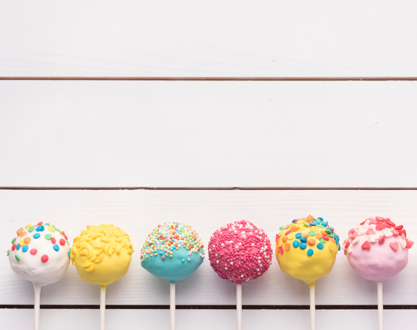 Six colorful cake pops with various sprinkles are lined up against a white wood-plank background.