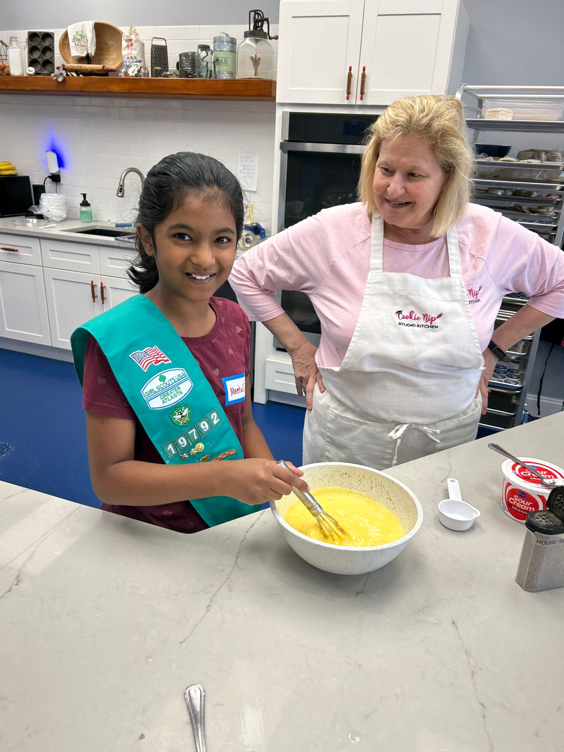 A girl wearing a sash and a woman in an apron stand in a kitchen; the girl uses a whisk to mix yellow batter in a bowl.
