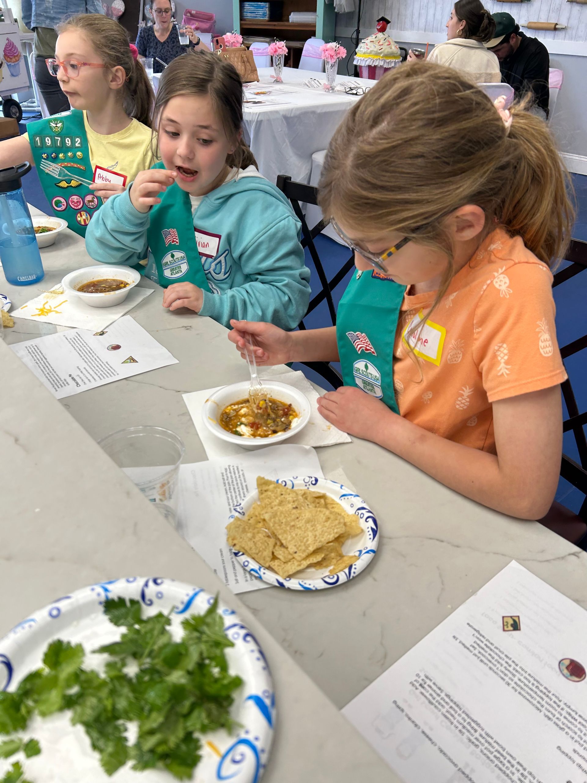 Three people in green sashes eat food from small bowls at a table set with crackers, cilantro, and instructional papers.