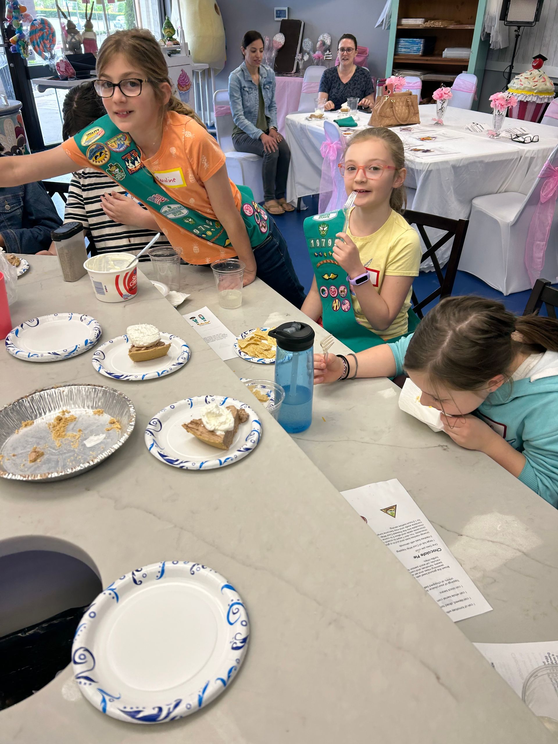 Participants in sashes eat slices of pie at a table in a brightly lit room decorated for an event.