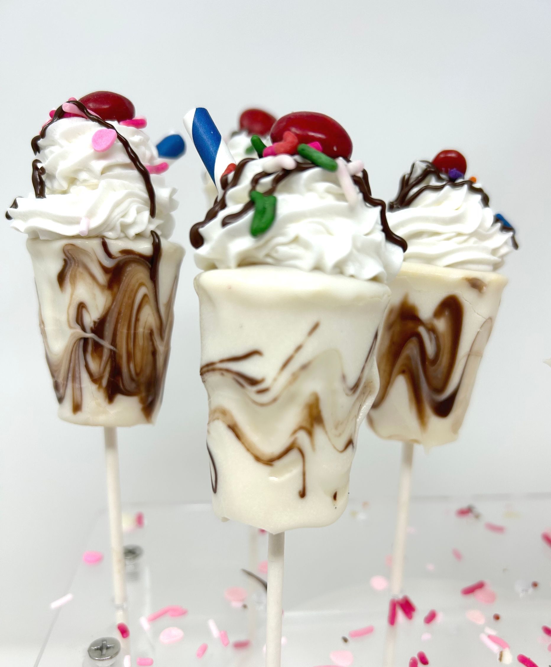Three milkshake-themed cake pops with white chocolate, brown drizzle, whipped frosting, sprinkles, and a cherry topper.