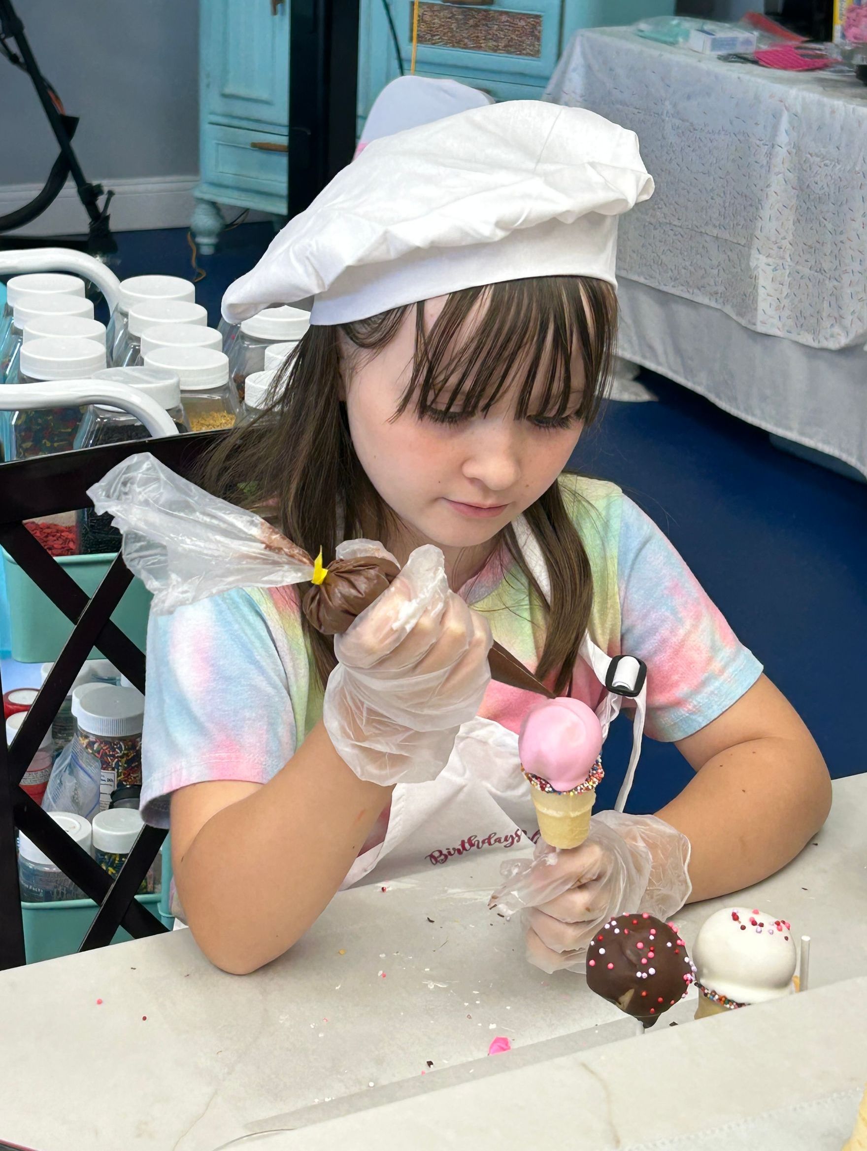 A person wearing a chef hat and gloves decorates small treats with frosting in a Cookie Nip Studio Kitchen workshop.