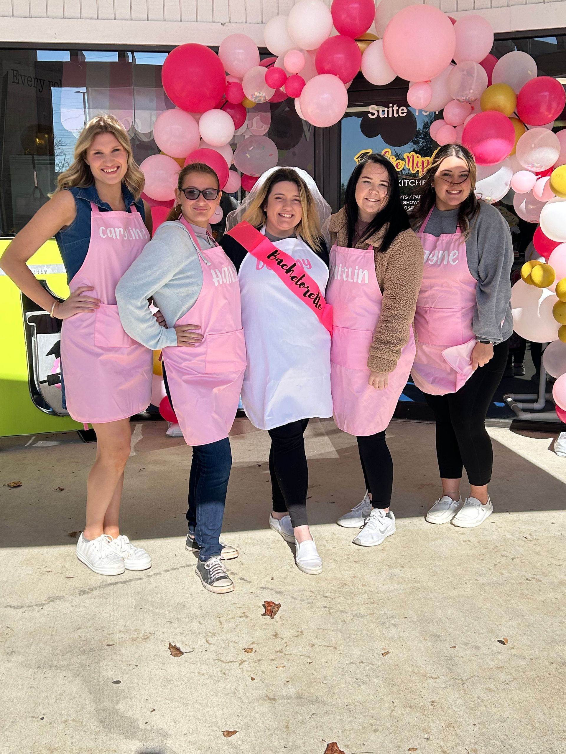 Five people stand smiling in front of a balloon arch, wearing matching pink aprons; the center person wears a bridal veil.