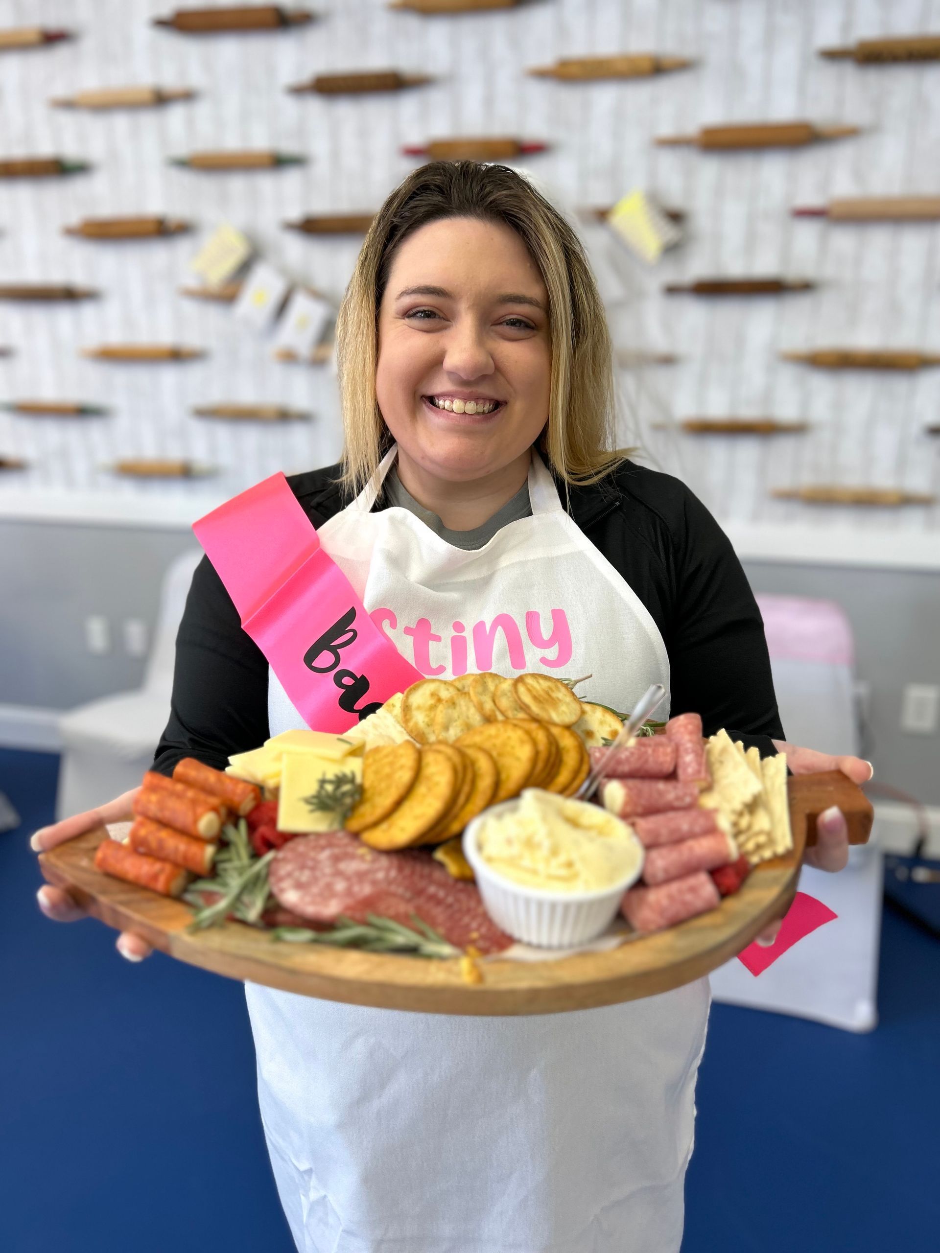 A person wearing an apron and pink sash holds a wooden charcuterie board of snacks in front of a wall with rolling pins.