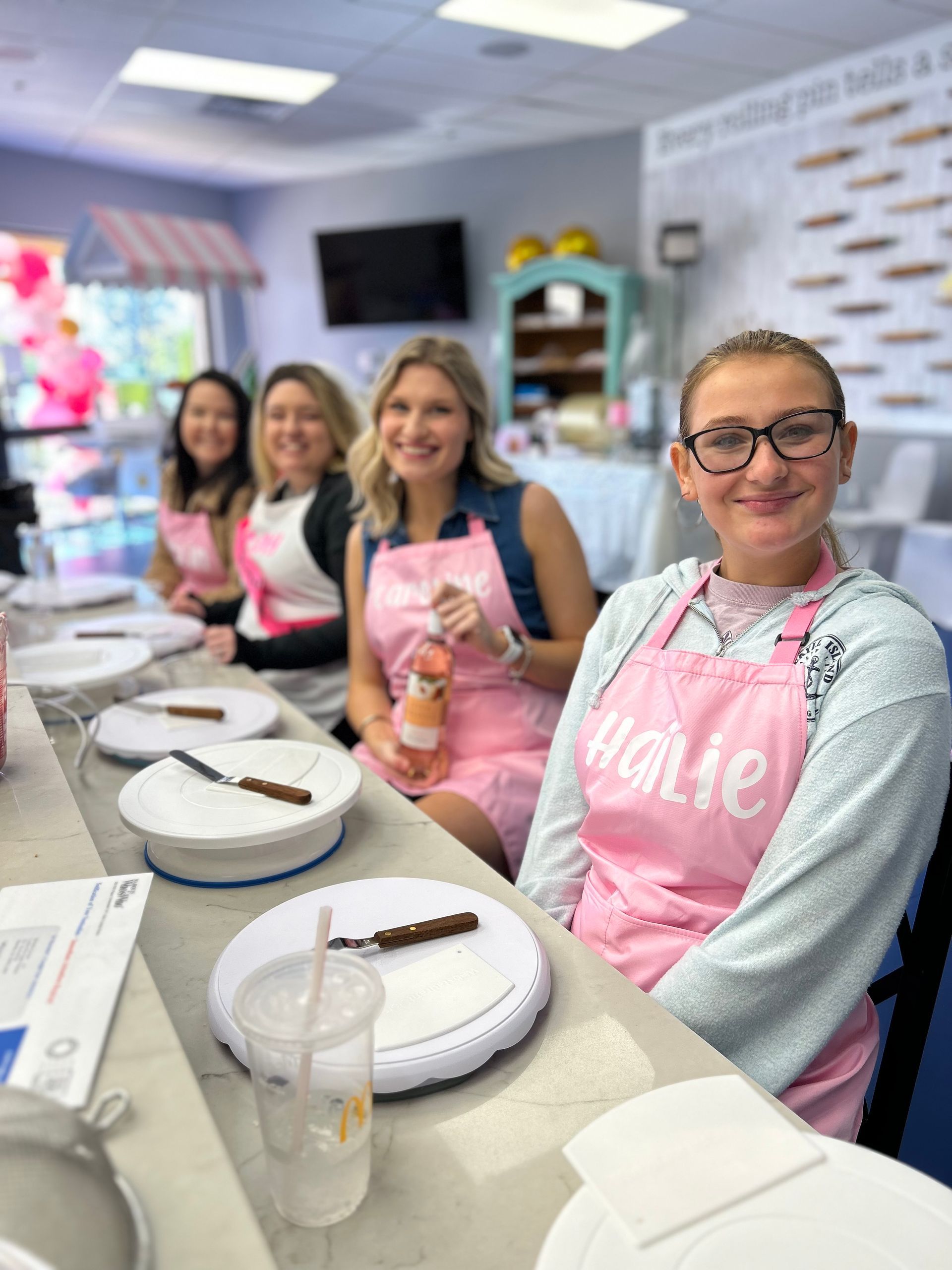 Four people in pink aprons sit at a table in a bright workshop, preparing to decorate cookies on white plates.