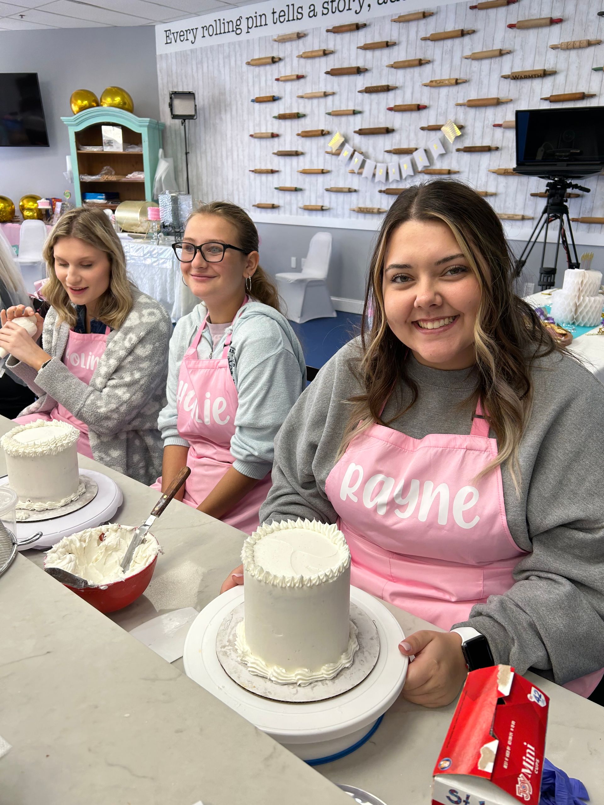 Three people in pink aprons sit at a counter, smiling while decorating small white cakes in a bright, decorated studio.