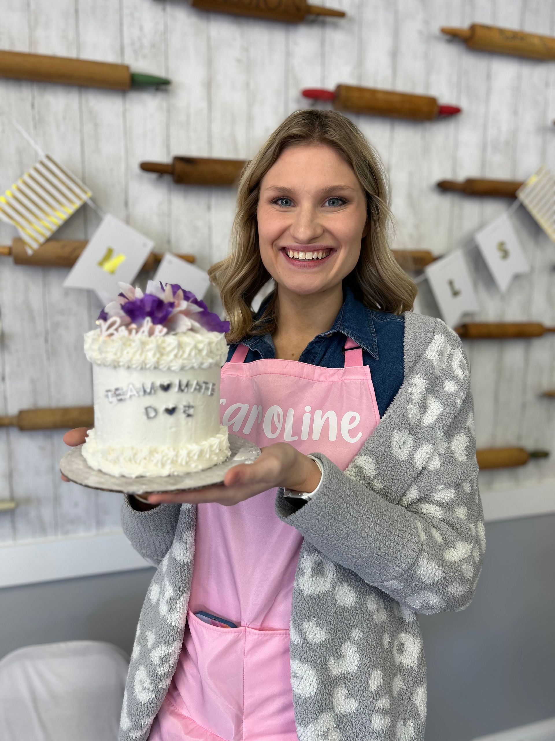 A smiling person wearing a pink apron and grey cardigan holds a small, white-frosted cake with purple accents.