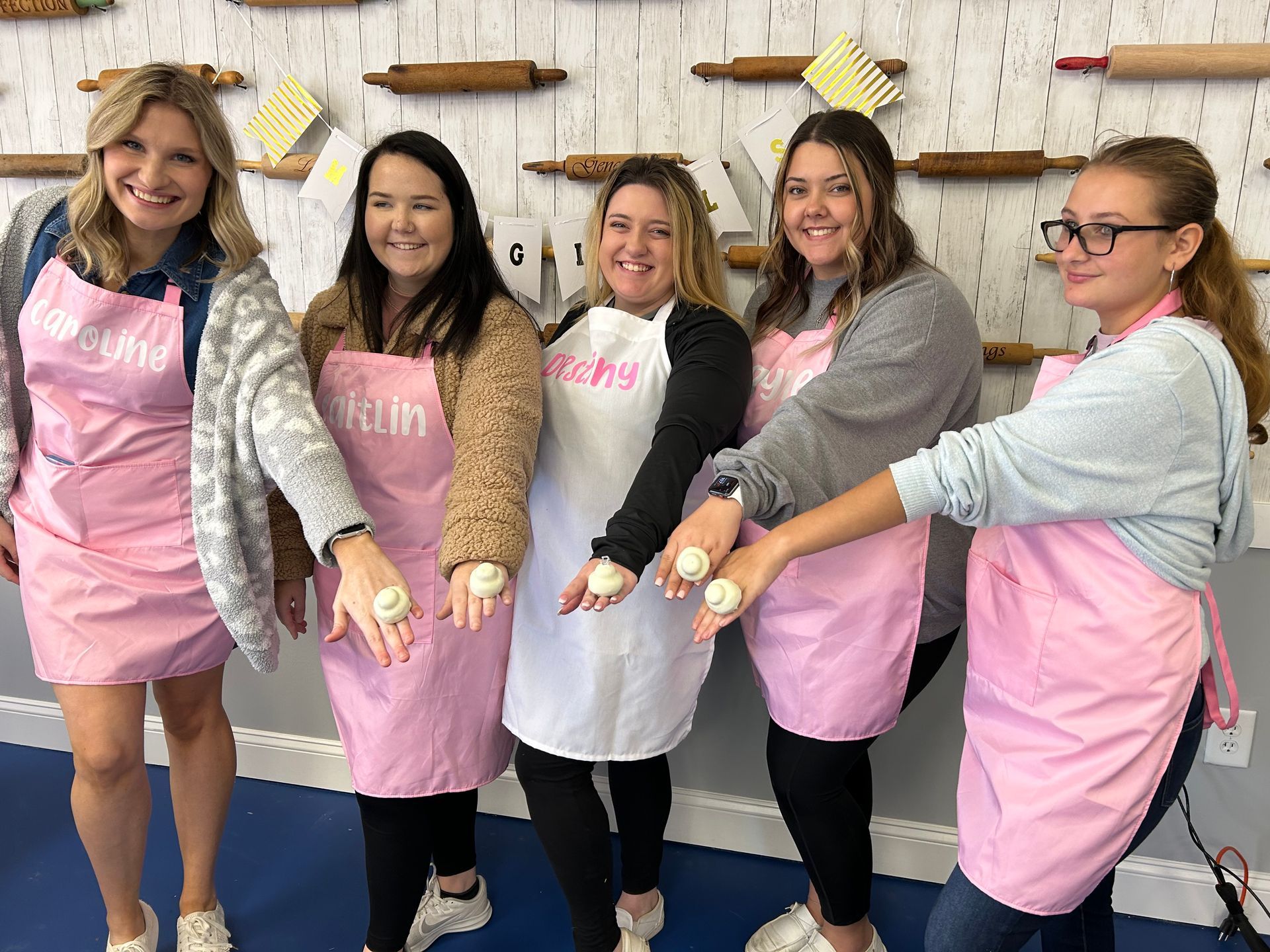 Five smiling people wearing pink aprons stand in a line against a wall of rolling pins, holding small cakes in their hands.