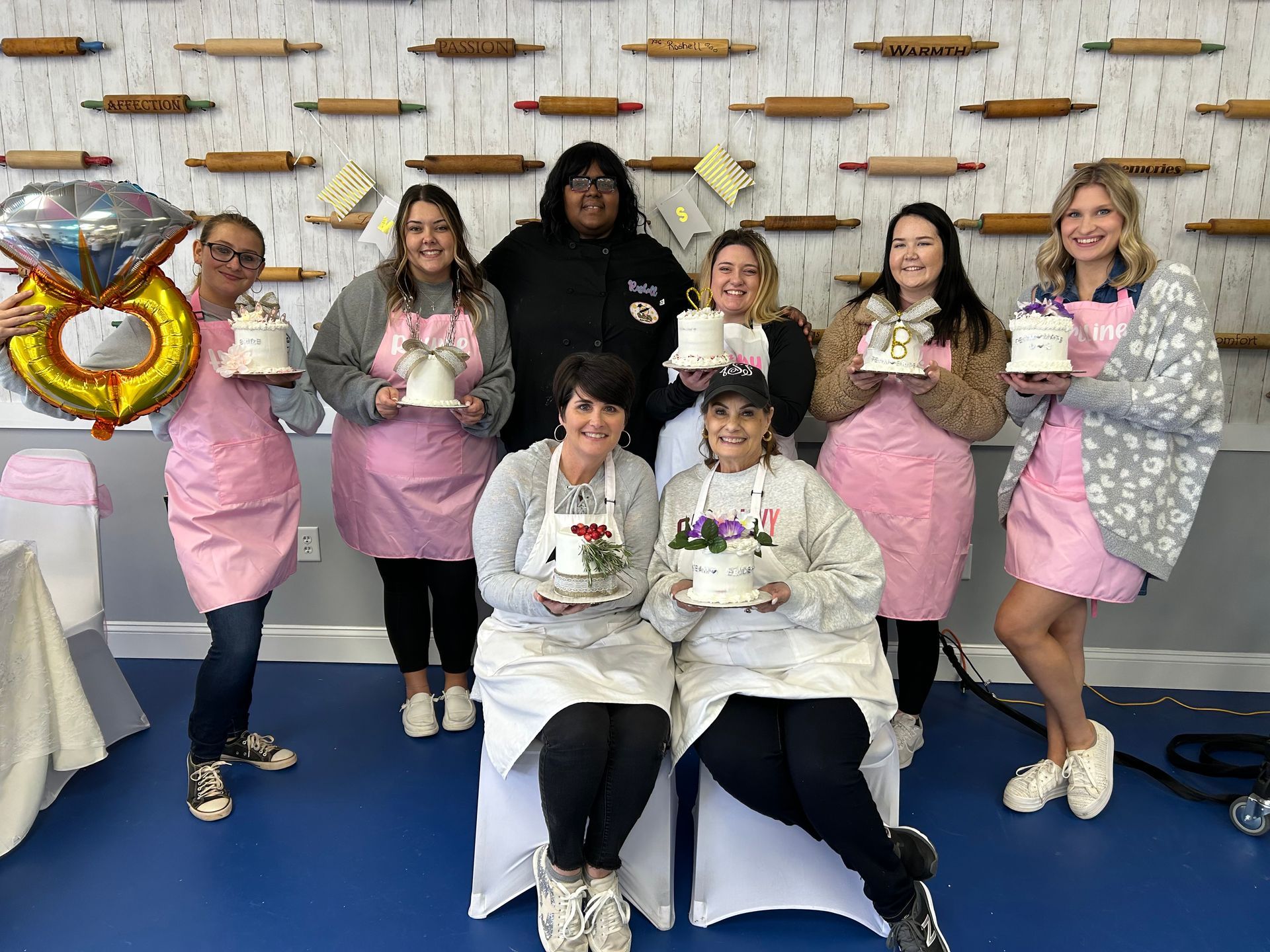 Group of people, mostly in pink aprons, holding decorated cakes, posing for a photo.