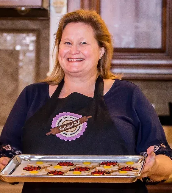 A person wearing a black apron holds a metal tray of decorated floral-shaped cookies in a kitchen.