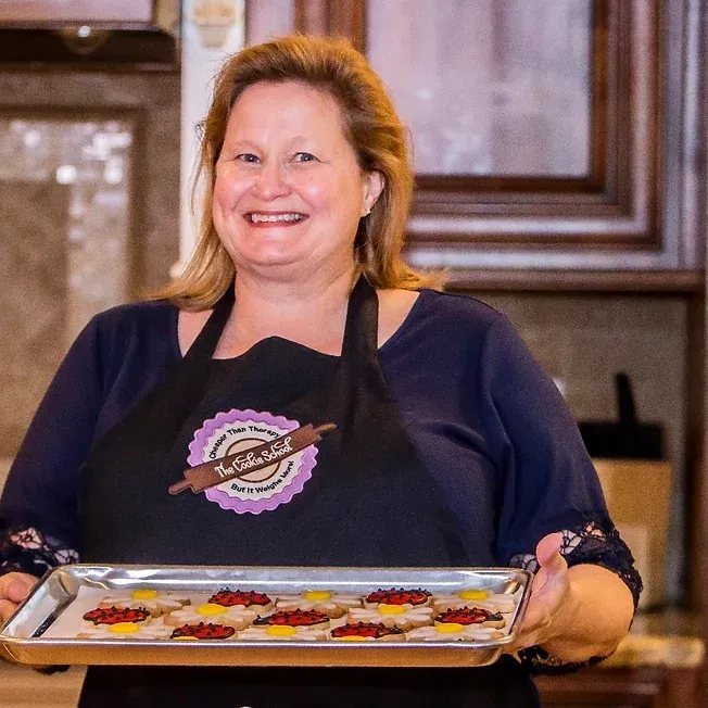 A smiling baker wearing a black apron holds a tray of colorful decorated flower cookies in a kitchen.