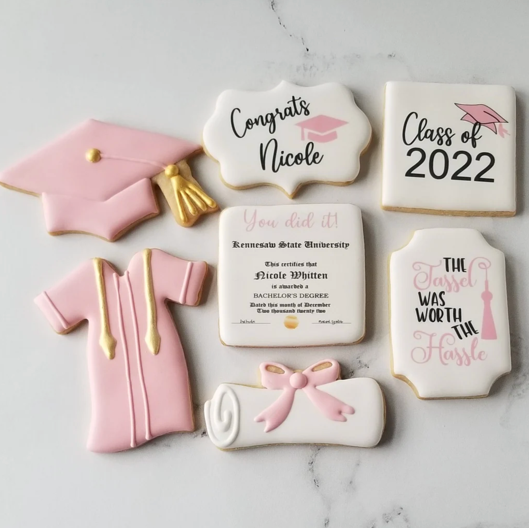 A collection of pink and white graduation-themed sugar cookies, including a cap, gown, diploma, and congratulatory text.
