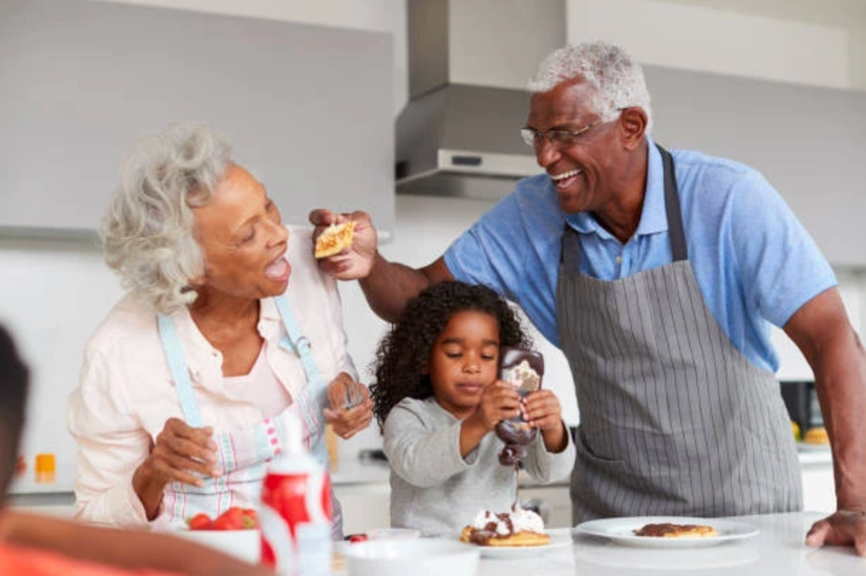 A child and two adults prepare and eat snacks together in a kitchen, smiling as the child decorates a small pancake.