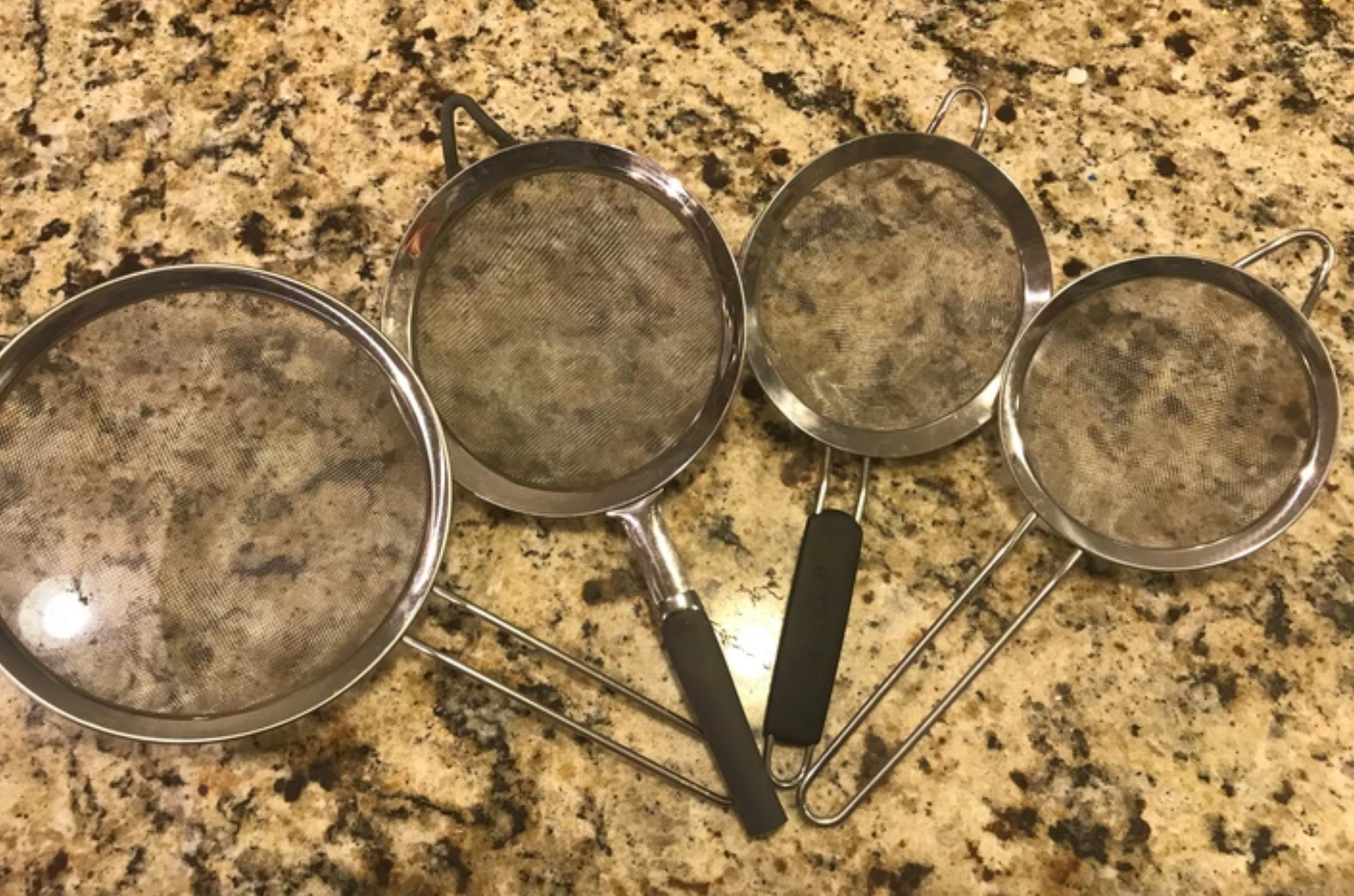 Four metal sieves of varying sizes on a speckled countertop.