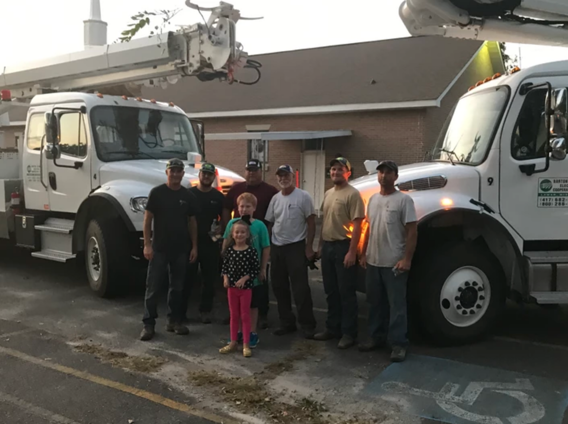 Group of people with utility trucks in front of a building. A young girl stands with them.