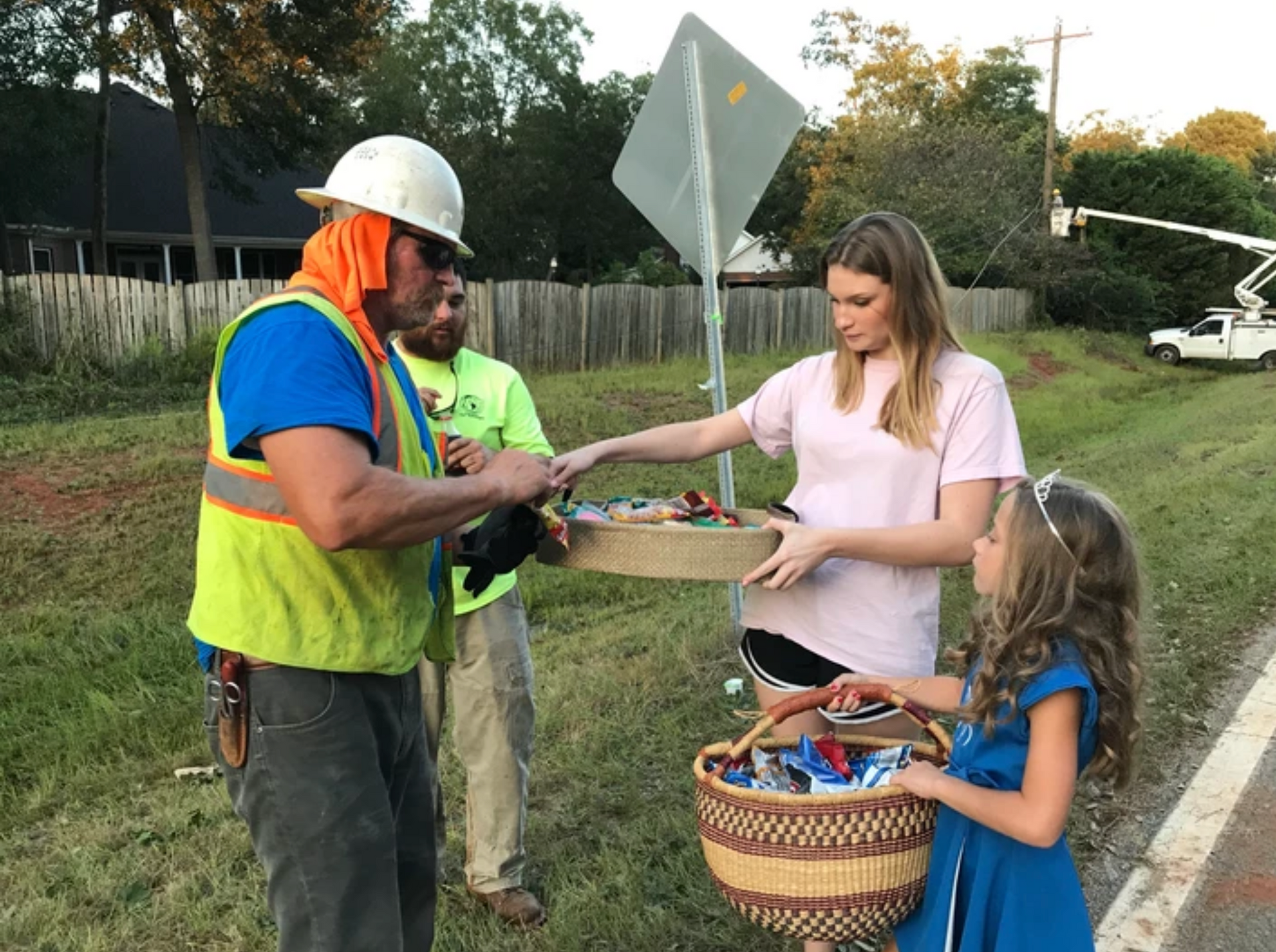 Utility workers and young girls offer treats by roadside, under a street sign, near a vehicle.