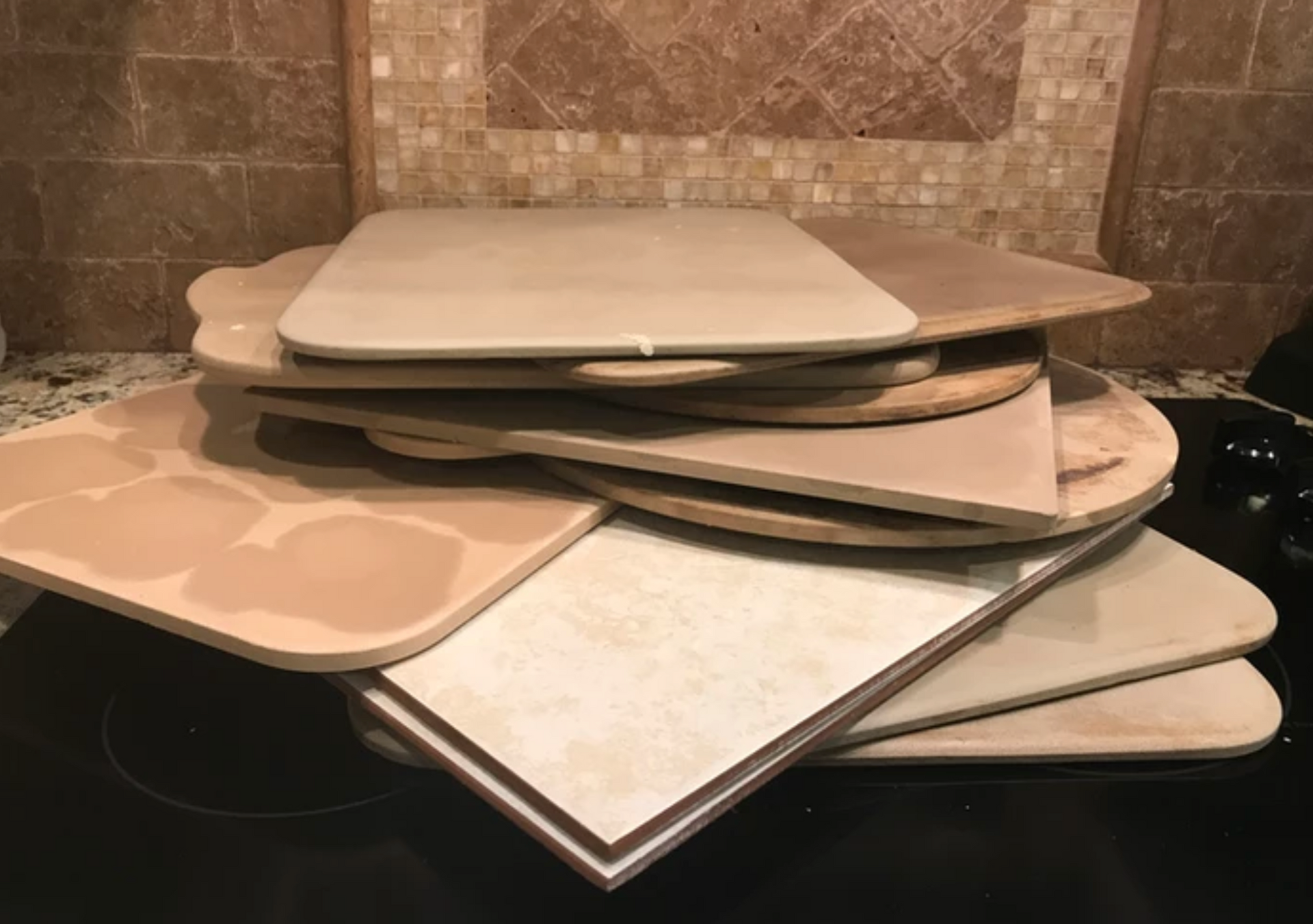 Pile of pizza stones and a ceramic tile on a stovetop, various shapes, beige and tan colors.