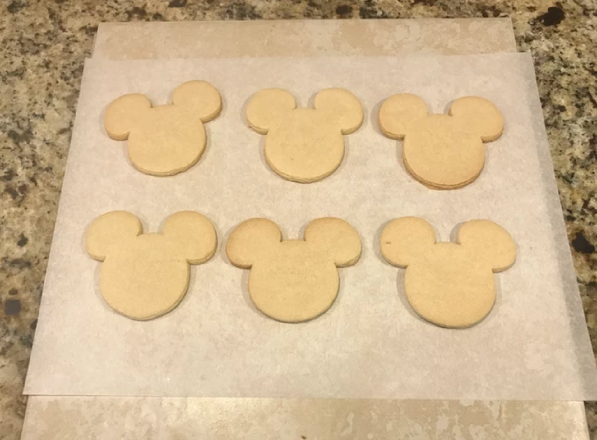 Six Mickey Mouse-shaped cookies on parchment paper, ready to be decorated.