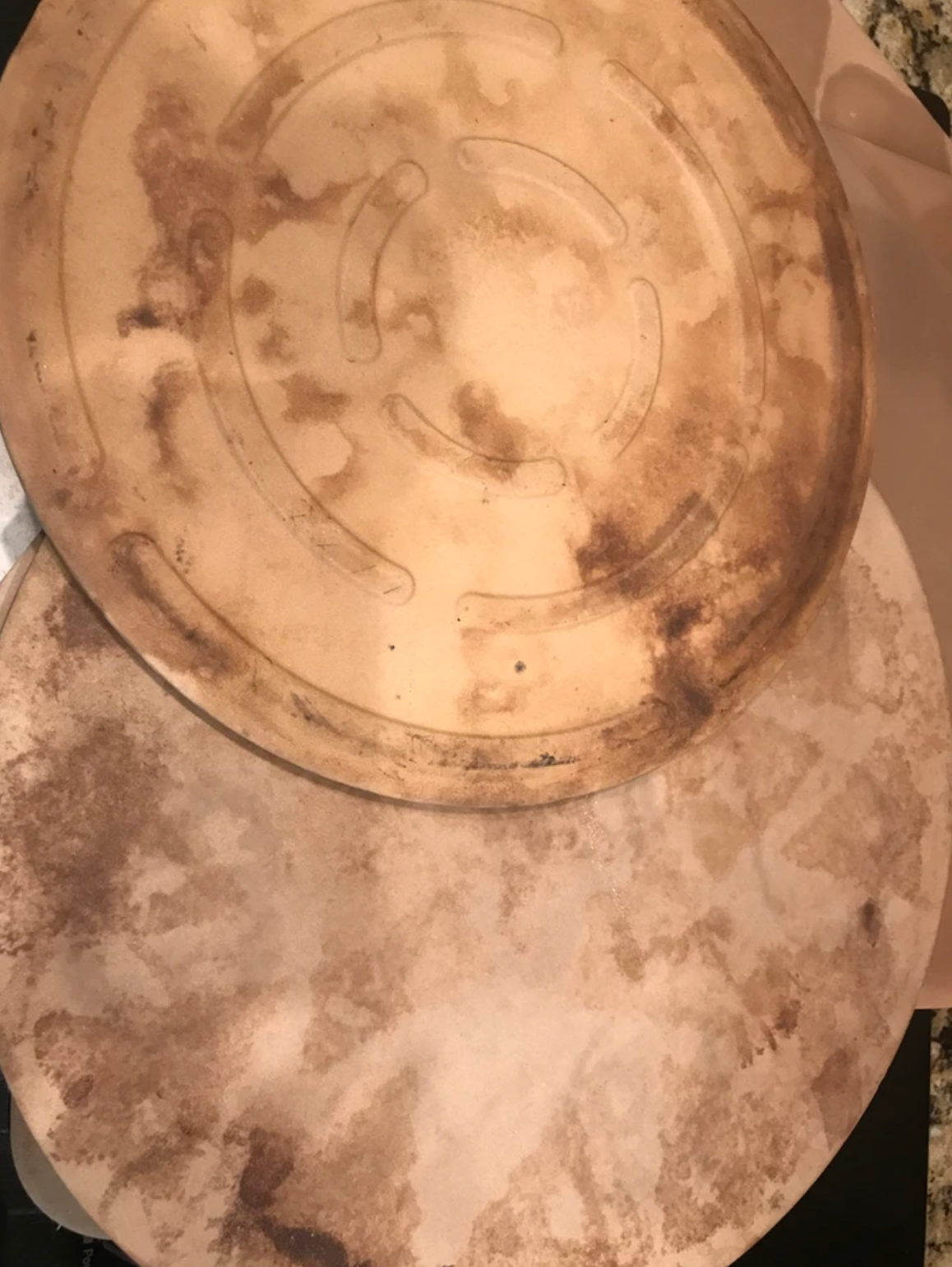 Two well-used, brown pizza stones with dark stains and circular grooves.