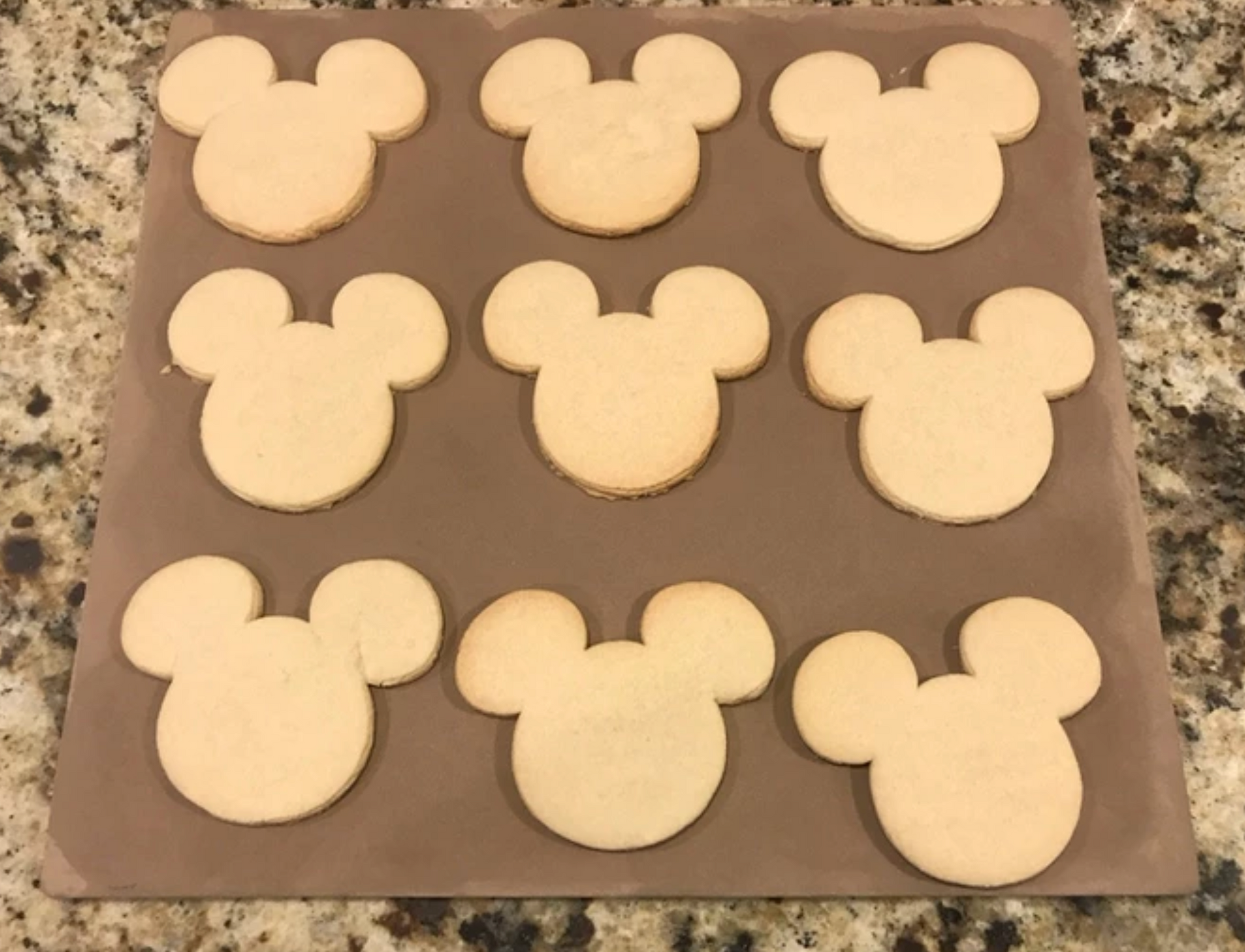 Nine Mickey Mouse-shaped cookies on a brown baking sheet.