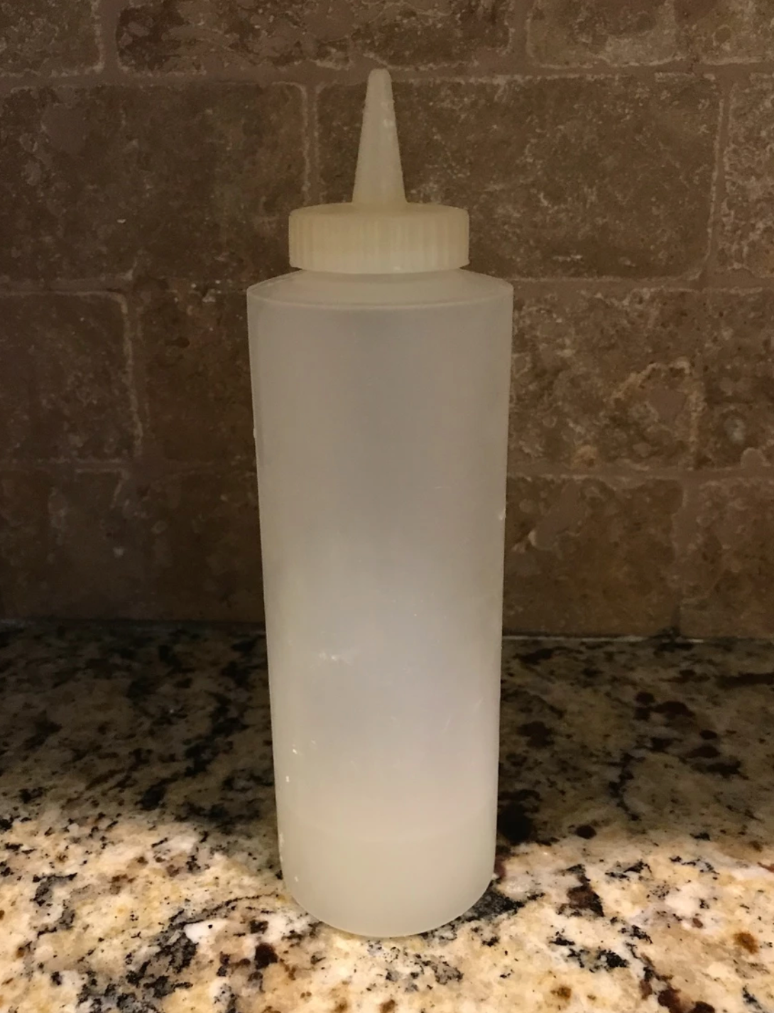 Clear plastic squeeze bottle with a pointed nozzle, resting on a speckled countertop.