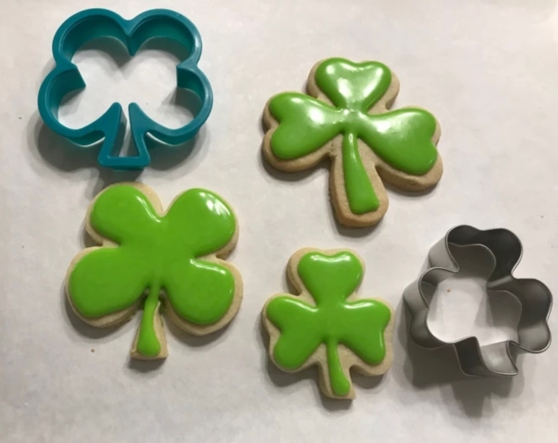 Shamrock cookies with green icing, next to blue and silver clover-shaped cookie cutters on white surface.