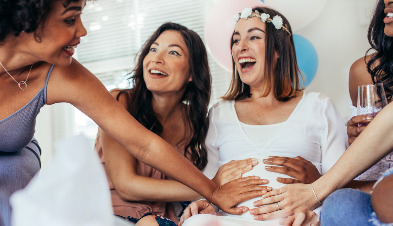 A group of friends gathered at a baby shower, smiling and touching a pregnant woman's belly.