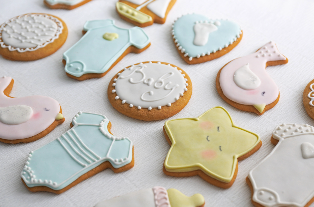 Cookies decorated with pastel-colored icing, shaped as baby-themed objects: onesie, bird, star, foot, bottle, and boat.