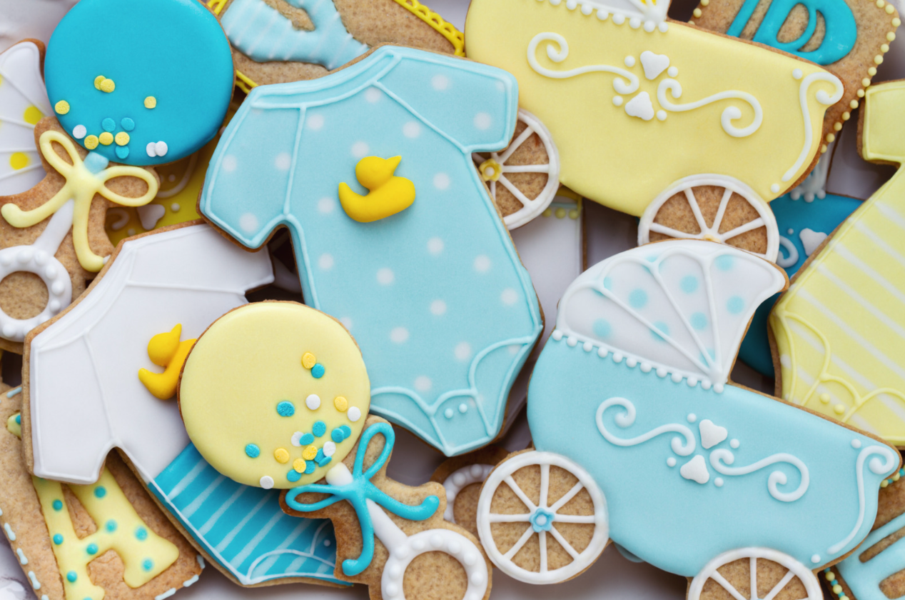 Decorative baby-themed sugar cookies, including ones shaped like onesies, rattles, and carriages in blue and yellow.