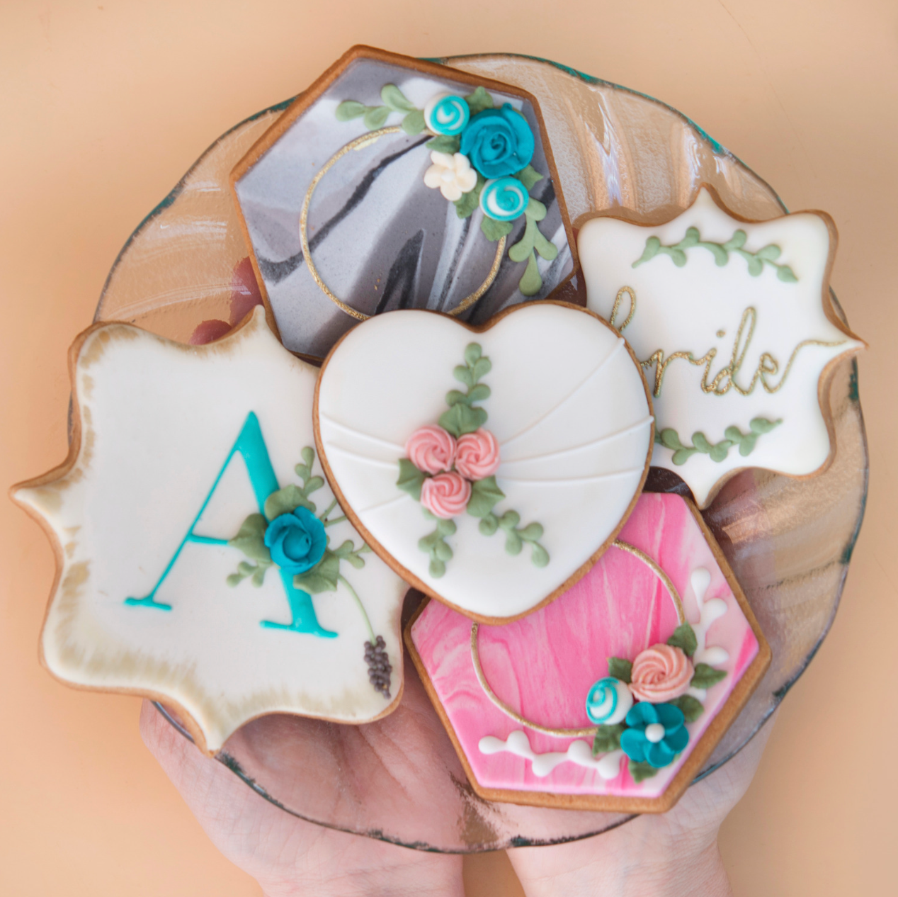 A person holding a glass plate with five decorative sugar cookies featuring floral designs, text, and marbled icing.