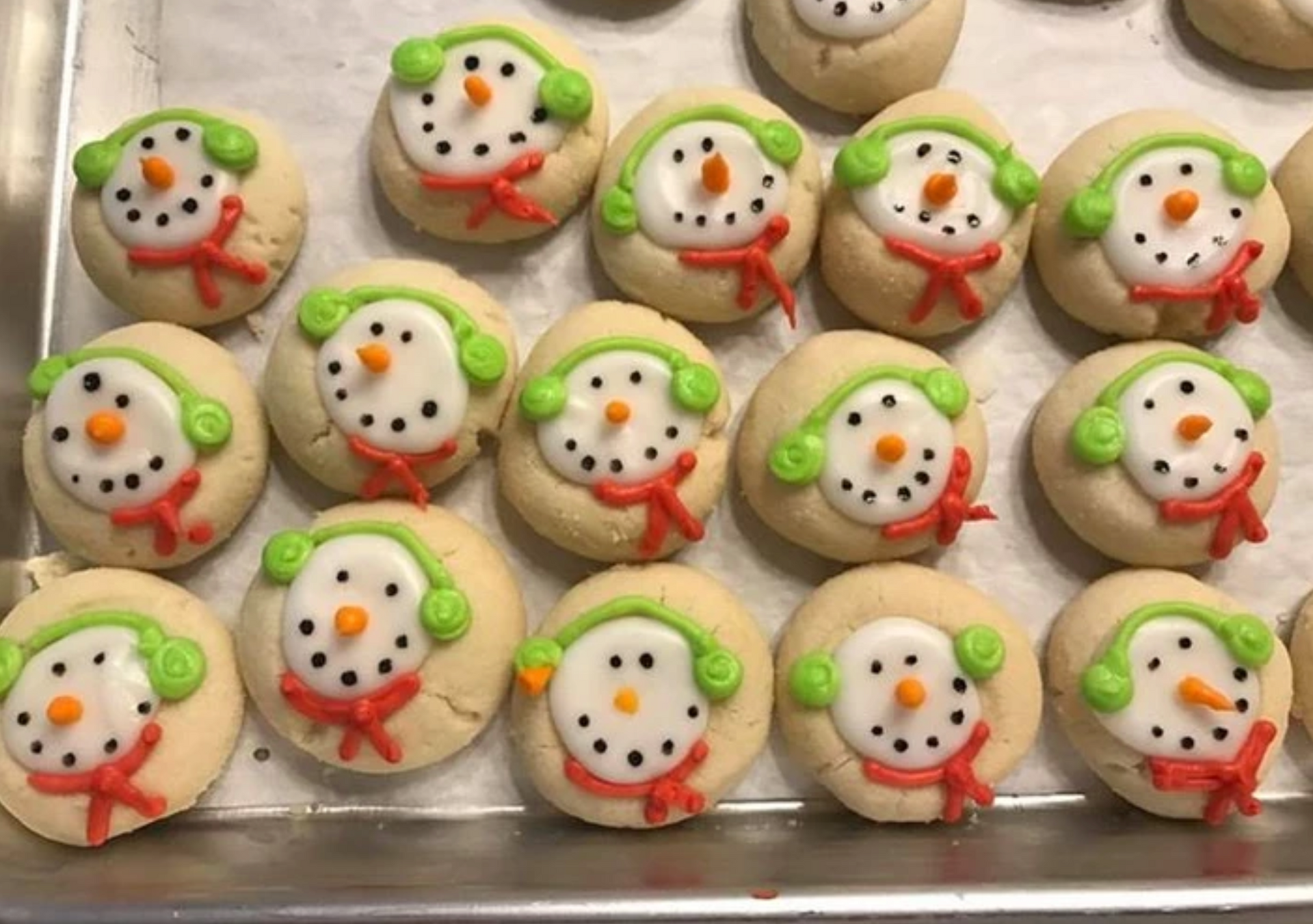Snowman-decorated sugar cookies on a baking sheet: white frosting, green earmuffs, red scarves, black dots for eyes.