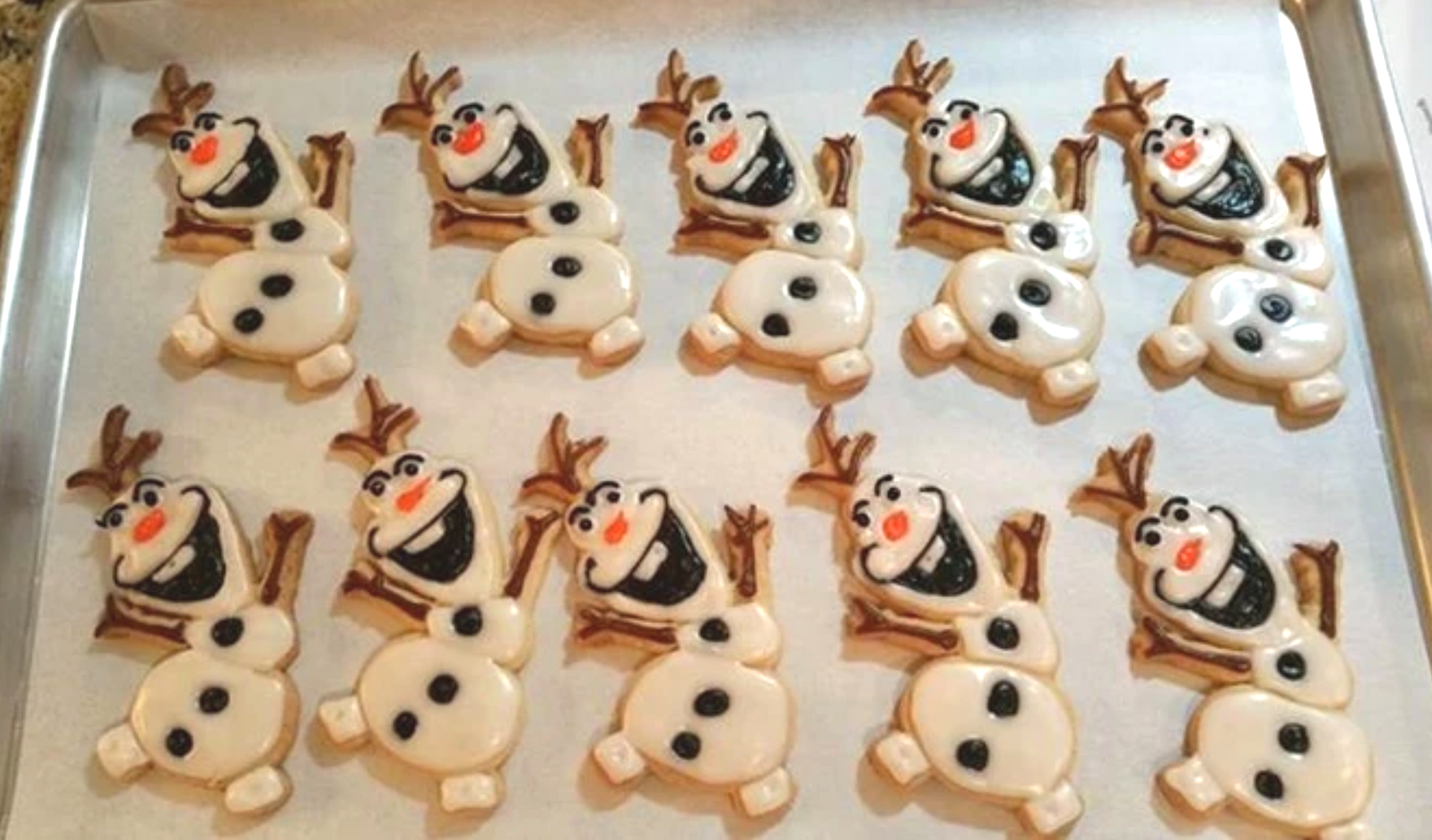 Olaf cookies with white icing, black details, and brown twig arms and hair, arranged on a baking sheet.