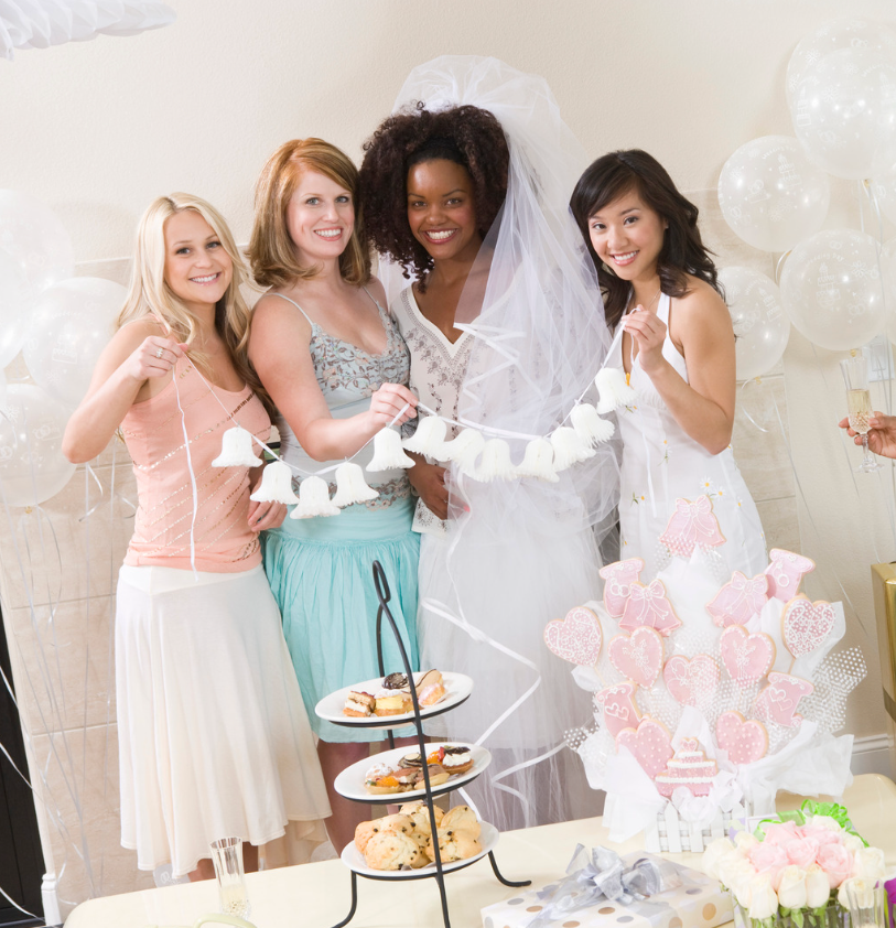 Four people smiling at a bridal shower, holding a bell banner in front of a tiered serving tray and pink heart decorations.
