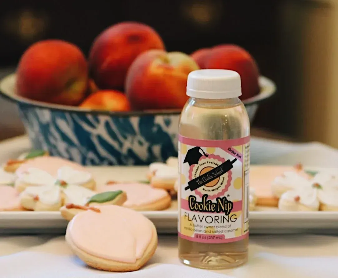 A bottle of Cookie Nip flavoring sits next to peach-shaped sugar cookies in front of a bowl of fresh peaches.