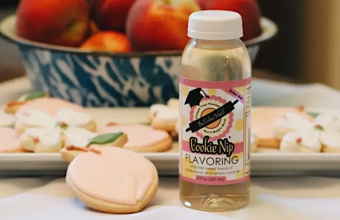 A bottle of Cookie Nip flavoring next to a plate of peach-shaped iced cookies, with a bowl of fresh peaches in the back.