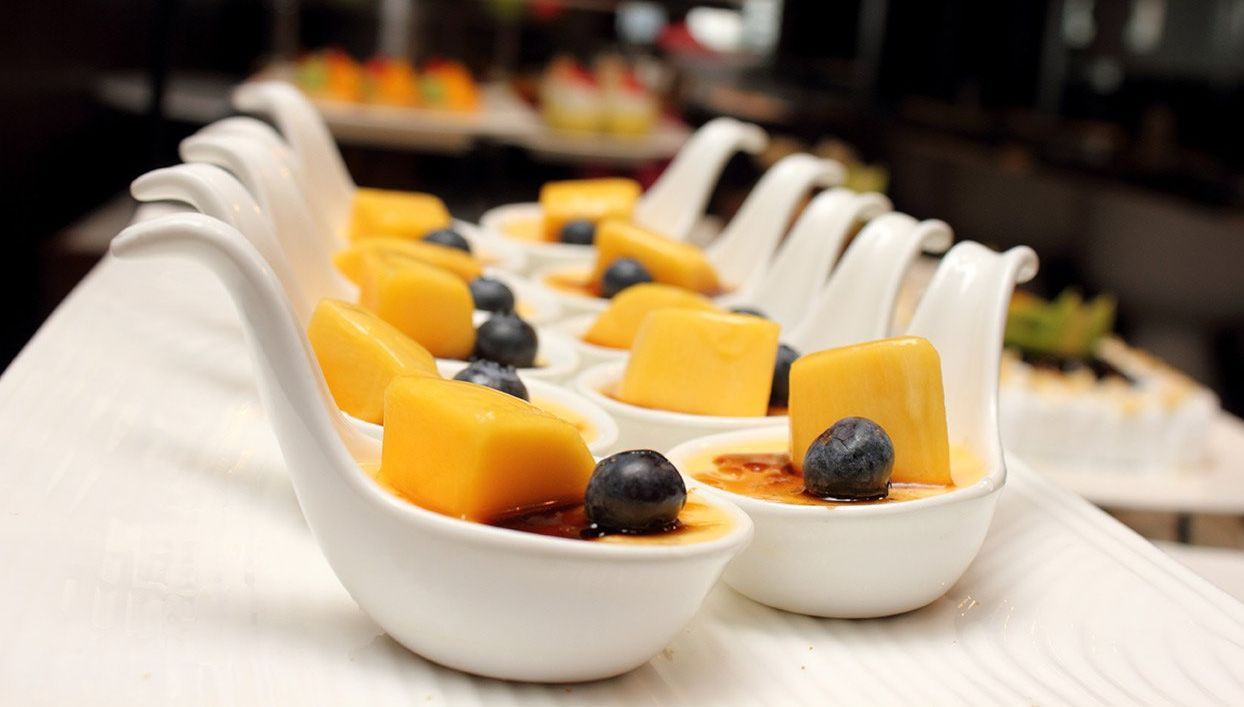 Small white spoons arranged in a row, each containing a custard dessert topped with a piece of mango and a blueberry.
