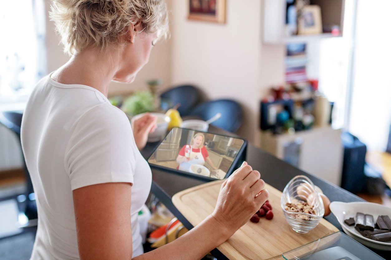 A person prepares food on a wooden board while watching a cooking tutorial on a tablet in a home kitchen.