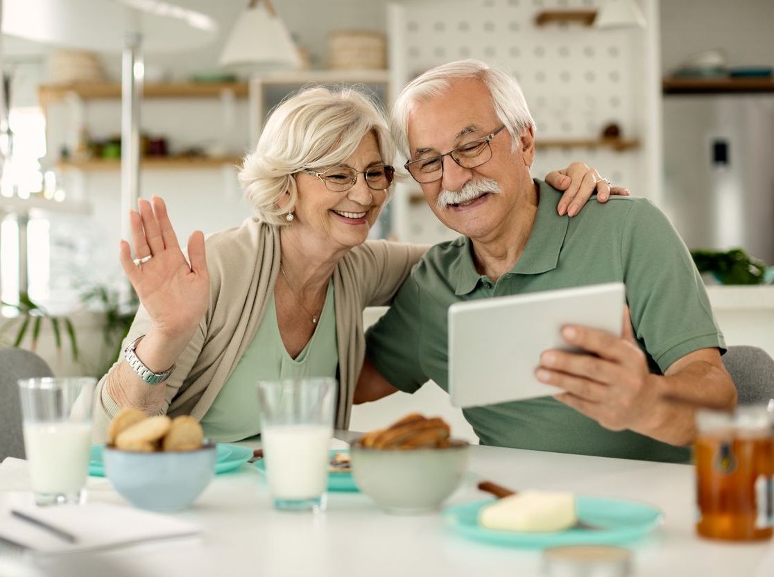 Two people sitting at a table with glasses of milk and snacks, smiling while having a video call on a tablet.