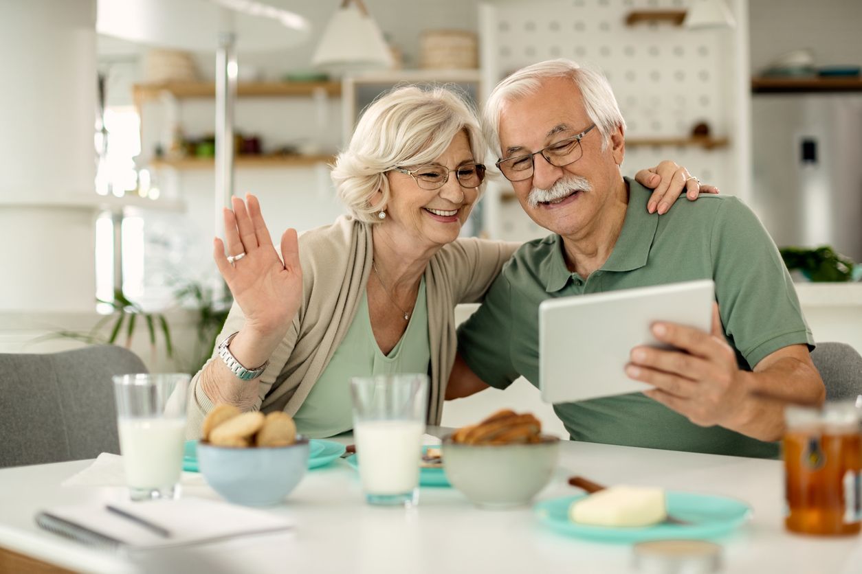 Two people at a kitchen table using a tablet to video call while waving, with glasses of milk and snacks nearby.