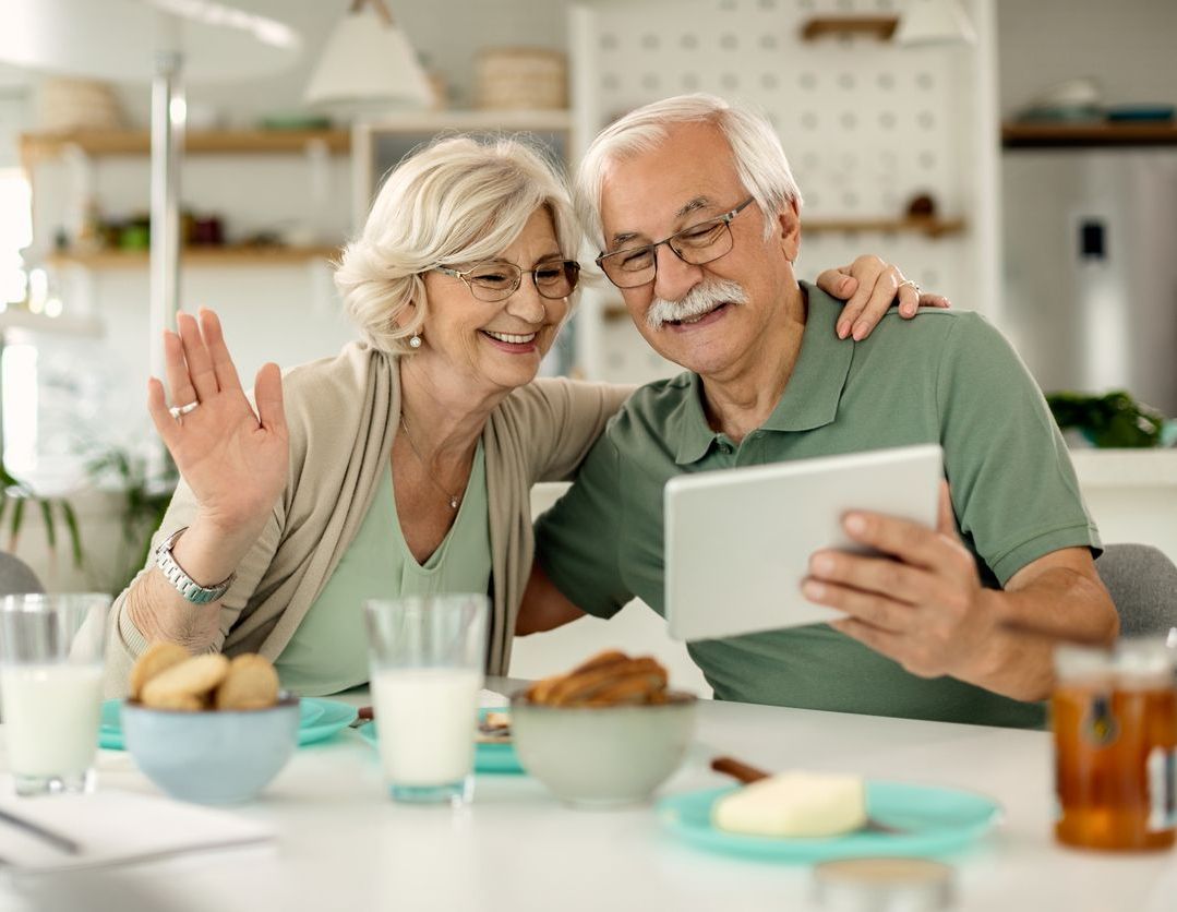 An older couple smiling while sitting at a kitchen table, using a tablet to have a video call.