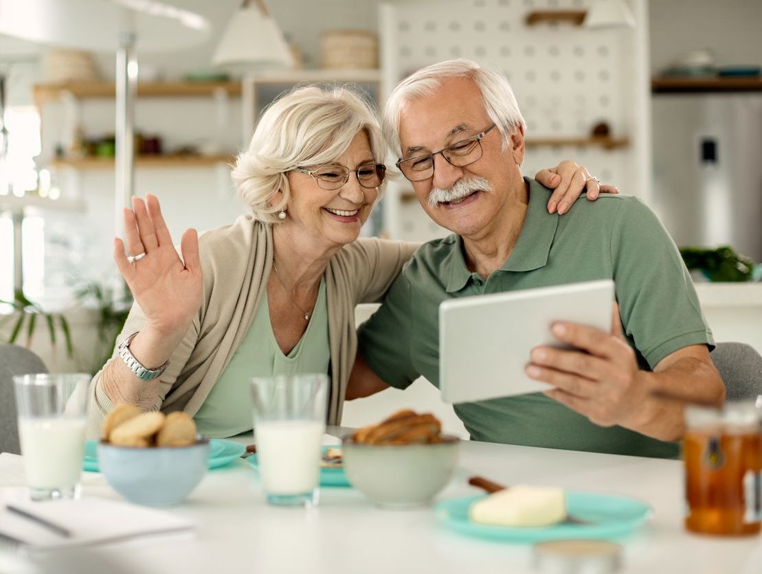 Two people sitting at a table together having a video call on a tablet, smiling and waving.
