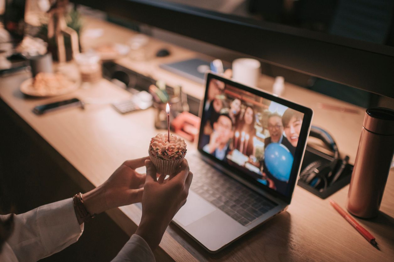A person holds a cupcake with a lit candle while participating in a virtual birthday video call on a laptop at a desk.