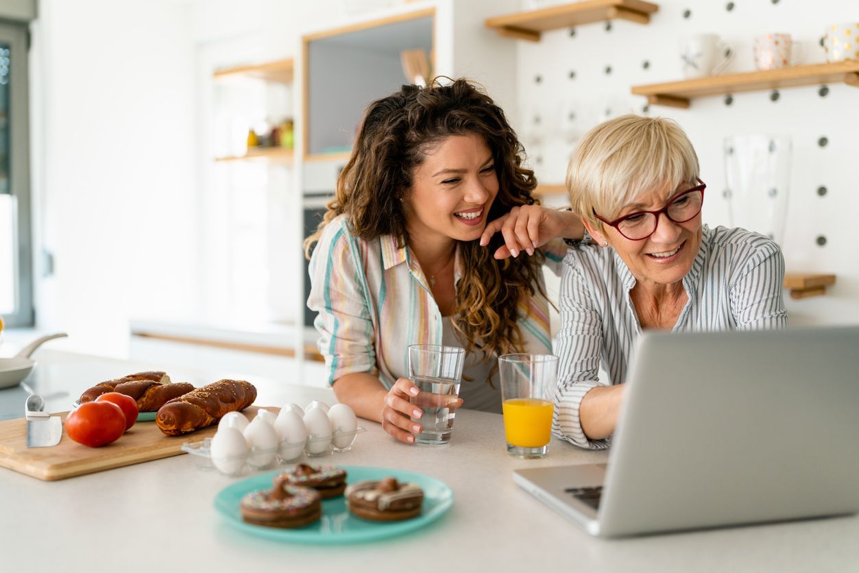Two people sitting at a kitchen counter, smiling while looking at a laptop, with pastries and orange juice nearby.
