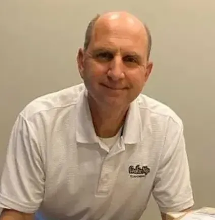 A person with a neutral expression wears a white polo shirt with a logo, positioned in front of a plain wall.