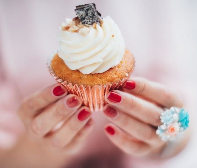 Hands with red-painted nails hold a cupcake topped with white frosting and a small chocolate piece, wearing a floral ring.
