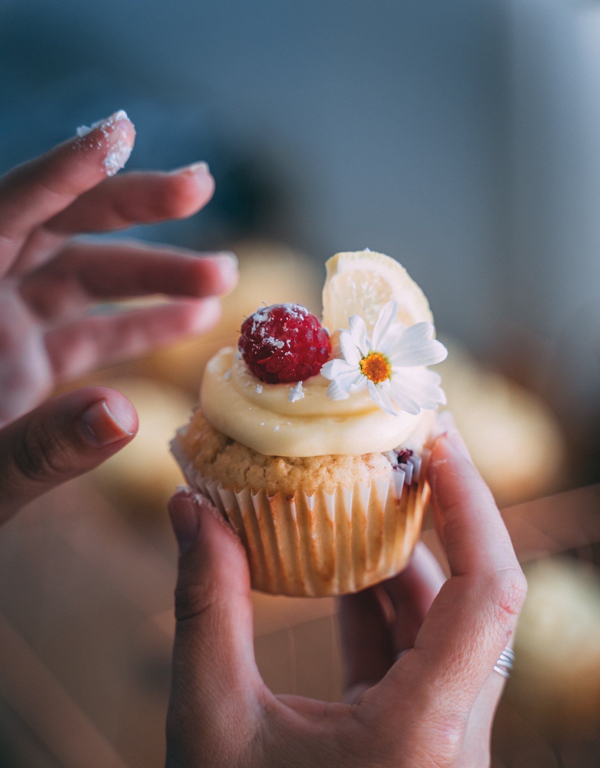A person holds a cupcake topped with frosting, a raspberry, a lemon slice, and a white flower, dusted with sugar.