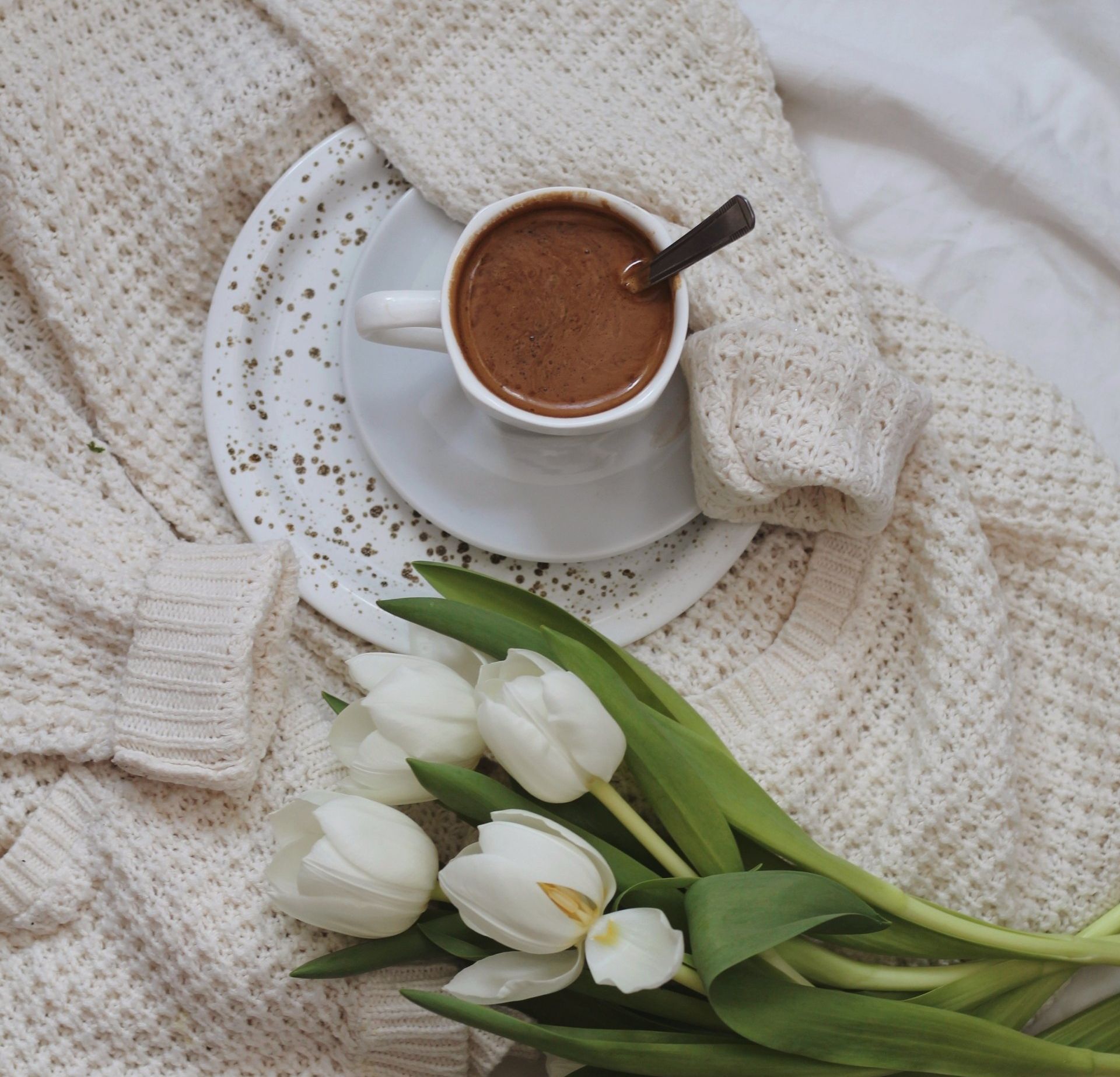 A cup of coffee on a speckled saucer atop a cozy cream-colored knit sweater, alongside a bouquet of white tulips.
