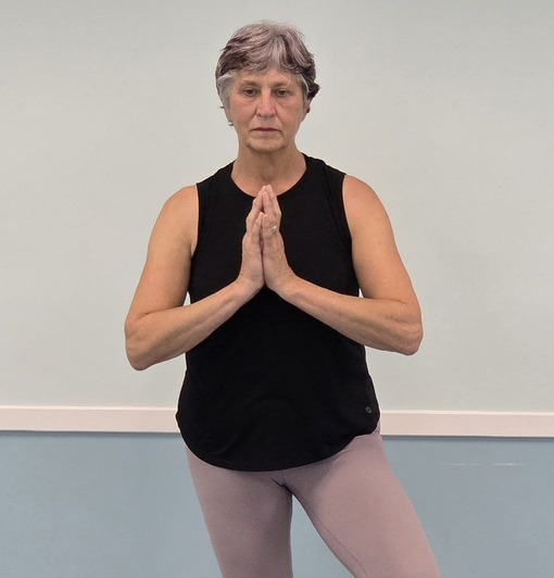 Person standing in yoga tree pose with hands in prayer, wearing black top and light leggings.